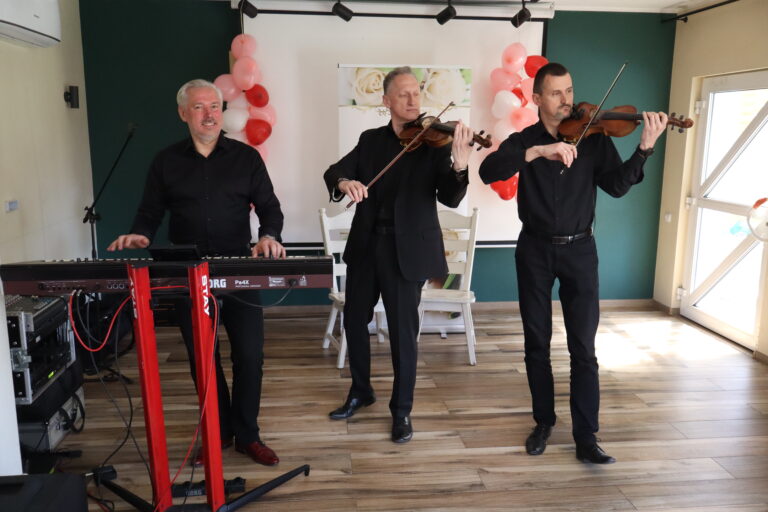 W sali okolicznościowej trzech muzyków wykonuje utwór na żywo. Po lewej stronie mężczyzna gra na keyboardzie, a obok niego dwóch instrumentalistów gra na skrzypcach. Wszyscy ubrani są elegancko na czarno. W tle widoczne są dekoracje z balonów w odcieniach bieli, różu i czerwieni oraz jasne wnętrze sali przygotowane na uroczystość.