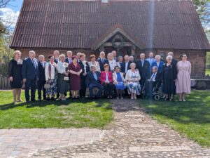 Grupa kilkudziesięciu osób, głównie seniorów i osób dorosłych, pozuje do wspólnego zdjęcia na trawniku przed zabytkowym drewnianym budynkiem z ciemnym dachem. Część uczestników stoi, a część siedzi na ławce ustawionej na środku. Kilka osób ma przypięte odznaczenia, co sugeruje oficjalny lub jubileuszowy charakter wydarzenia. Na pierwszym planie widoczna jest kamienna ścieżka prowadząca do budynku, a całość oświetla słoneczna pogoda