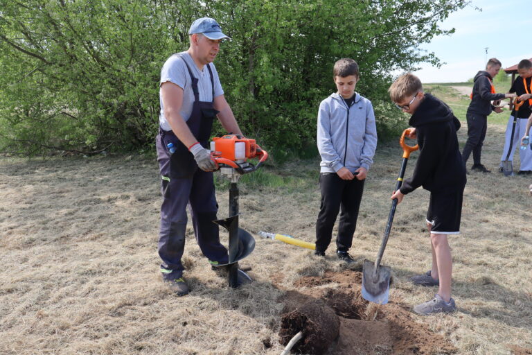 Mężczyzna z wiertnicą spalinową i dwaj chłopcy z łopatą przygotowują dół do posadzenia drzewa na trawiastym terenie.