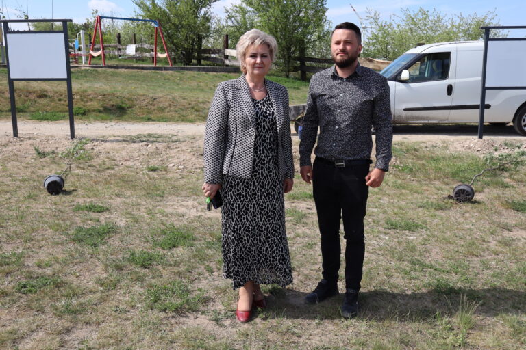 Kobieta w żakiecie i mężczyzna w koszuli pozują na zewnątrz; w tle biały furgonetka i plac zabaw.