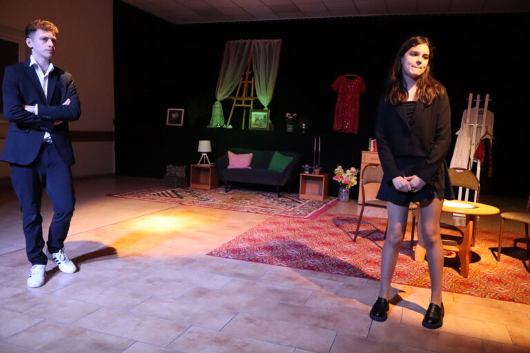 Dwoje młodych aktorów na scenie podczas spektaklu teatralnego — chłopiec w garniturze i dziewczyna w czarnej sukience na tle scenografii pokoju