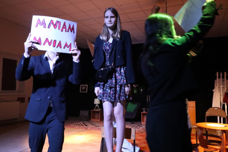 Scena spektaklu teatralnego — aktor trzyma tabliczkę z napisem "Mniam Mniam", dziewczyna w kwiecistej sukience stoi na krześle