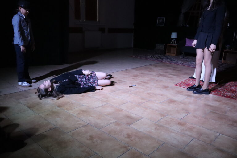 Dramatyczna scena spektaklu teatralnego — aktorka leży na podłodze, pozostałe osoby stoją wokół niej w scenicznym oświetleniu