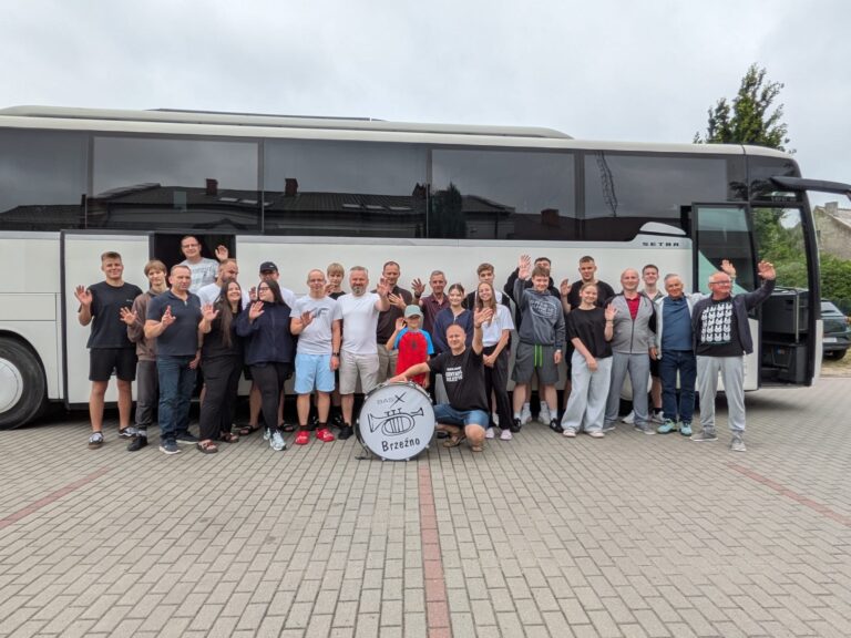 Grupa osób machających przed białym autokarem, z bębnem z napisem Brzeźno na pierwszym planie