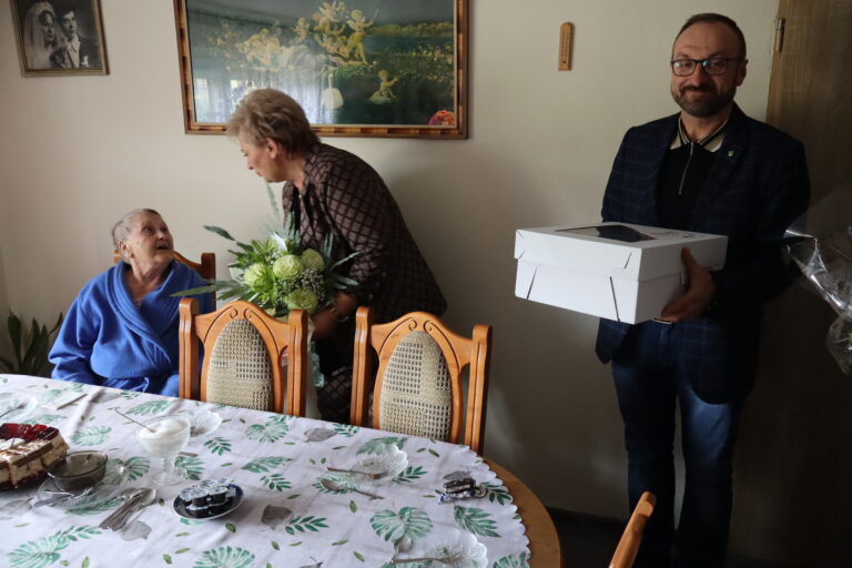 Kobieta wręcza kwiaty starszej pani siedzącej przy stole, obok stoi mężczyzna trzymający pudełko z tortem