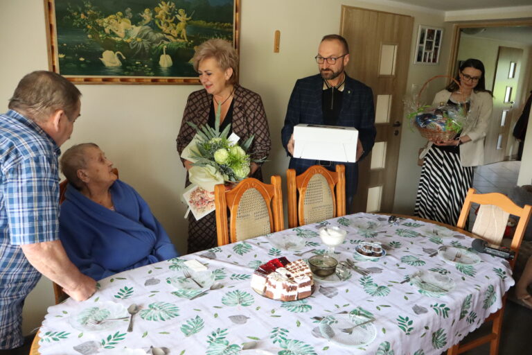 Goście wręczają kwiaty i prezenty starszej kobiecie siedzącej przy odświętnie nakrytym stole