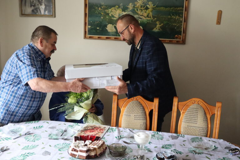 Dwóch mężczyzn wręcza pudełko z tortem osobie siedzącej przy stole z kwiatami i poczęstunkiem