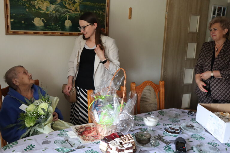 Starsza kobieta z bukietem kwiatów siedzi przy stole w otoczeniu dwóch kobiet podczas domowej uroczystości