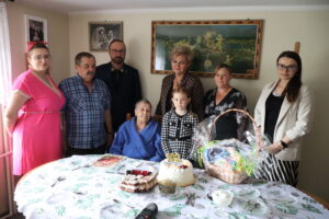 Starsza kobieta świętująca 102. urodziny w gronie rodziny przy stole z tortem i koszem upominkowym
