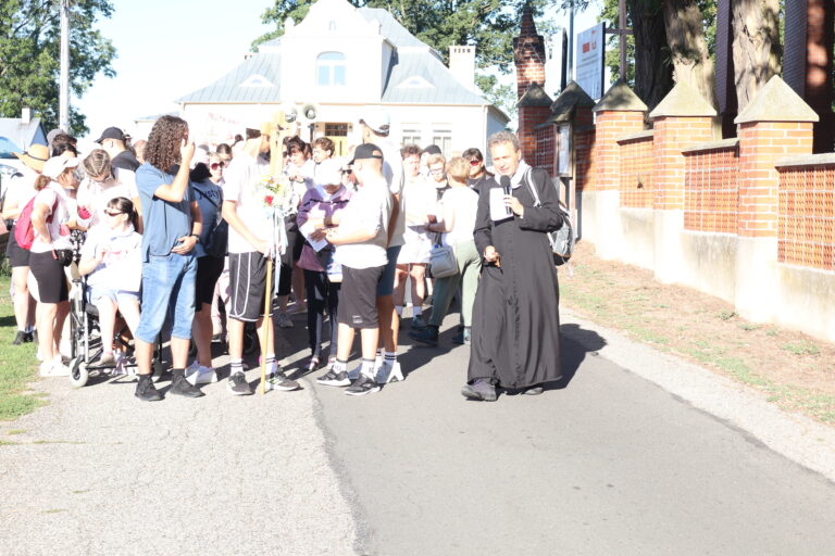 Ksiądz z mikrofonem prowadzi grupę pielgrzymów, w tym osobę na wózku i chłopaka niosącego krzyż procesyjny