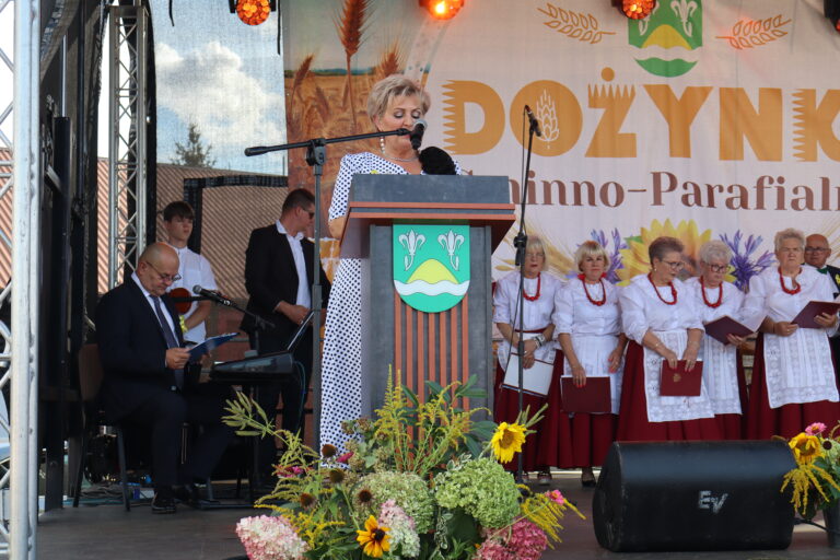 Kobieta przemawiająca przy mównicy podczas dożynek, w tle chór w tradycyjnych strojach ludowych