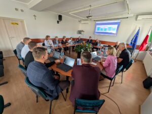 Osoby obradujące przy dużym stole z laptopami w sali konferencyjnej z wyświetloną prezentacją na ekranie