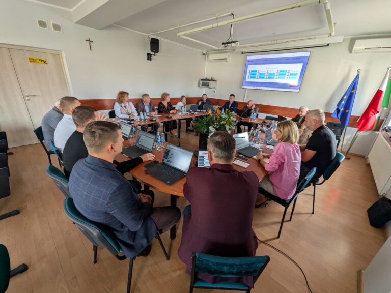 Osoby obradujące przy dużym stole z laptopami w sali konferencyjnej z wyświetloną prezentacją na ekranie