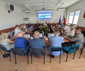 Grupa osób podczas obrad przy owalnym stole w sali z polskim godłem i flagami