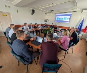 Osoby obradujące przy dużym stole z laptopami w sali konferencyjnej z wyświetloną prezentacją na ekranie