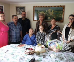 Starsza kobieta świętująca 102. urodziny w gronie rodziny przy stole z tortem i koszem upominkowym