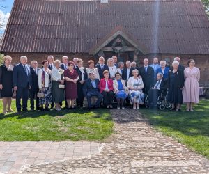 Grupa kilkudziesięciu osób, głównie seniorów i osób dorosłych, pozuje do wspólnego zdjęcia na trawniku przed zabytkowym drewnianym budynkiem z ciemnym dachem. Część uczestników stoi, a część siedzi na ławce ustawionej na środku. Kilka osób ma przypięte odznaczenia, co sugeruje oficjalny lub jubileuszowy charakter wydarzenia. Na pierwszym planie widoczna jest kamienna ścieżka prowadząca do budynku, a całość oświetla słoneczna pogoda